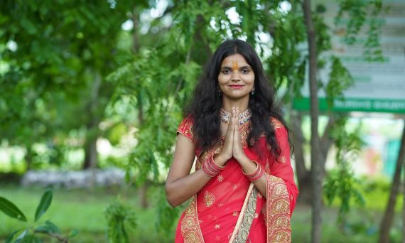 A young woman in a bright red saree standing outdoors, smiling gracefully with hands in a traditional pose.