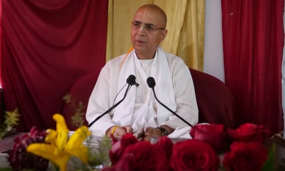Elderly man in white robes delivers a speech indoors with floral decorations.